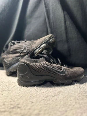 Svarta Nike Vapormax sneakers - Säljer ett par svarta Nike Vapormax sneakers med stickad ovandel och synlig luftdämpning i sulan. Skorna har snörning och en sportig, modern look med diskret Swoosh på sidan. Perfekta för dig som gillar stilrena och bekväma sneakers.