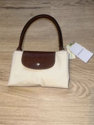 Beige handväska från Longchamp - Stilren beige handväska från Longchamp med bruna skinnhandtag och lock i brunt skinn. Väskan har en klassisk form och stängs med tryckknapp. Perfekt för dig som gillar tidlös design och vill ha en väska som funkar till det mesta. Den är helt ny och aldrig använd. Säljer då jag råkade köpa fel storlek!