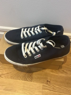 Marinblå sneakers från Helly Hansen - Snygga marinblå sneakers från Helly Hansen med vit sula och vita snören. Skorna har en klassisk låg profil och är tillverkade i textilmaterial. Helly Hansen-logga på sidan ger en sportig touch. Perfekta för en avslappnad och stilren look.