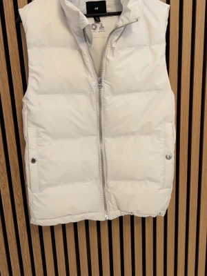 Vit dunväst från H&M XS - Säljer en vit dunväst från H&M i storlek XS. Västen har hög krage, dragkedja framtill och två fickor med tryckknappar. Materialet är vind- och vattenavvisande syntet, perfekt för kyliga dagar. Enkel och stilren design som passar till det mesta.