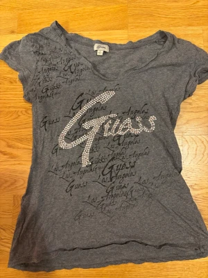 Grå t-shirt med strass från Guess - Snygg grå t-shirt från Guess med korta ärmar och coolt tryck med texten 'Guess' och 'Los Angeles' över hela framsidan. Stor logga i strass framtill som ger extra bling. Mjuk och tunn,  perfekt till jeans eller shorts.