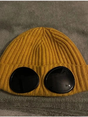 Gul ribbstickad mössa med glasögon - Säljer en gul ribbstickad mössa med inbyggda svarta glasögon framtill. Mössan har en cool och unik design som sticker ut och passar perfekt för dig som vill ha något annorlunda. Perfekt accessoar för höst och vinter.