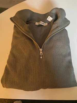 Grön stickad halfzip från Only & Sons - Snygg Grön stickad tröja från Only & Sons med ribbad struktur och halv dragkedja framtill. Tröjan har hög krage och långa ärmar, perfekt för lager på lager. Materialet är mjukt och stretchigt för extra komfort. Helt ny aldrig använd