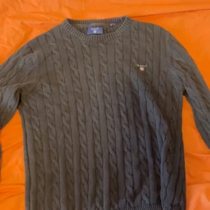 Mörkgrå kabelstickad tröja Gant - Snygg mörkgrå kabelstickad tröja från Gant med rund halsringning och diskret logga broderad på bröstet. Tröjan har långa ärmar och ribbade muddar vid ärmslut och nederkant. Perfekt för lager på lager och klassisk stil.