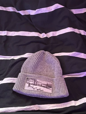 Grå ribbstickad mössa från Revenite - Säljer en grå ribbstickad mössa från Revenite med uppvikt kant och en snygg patch framtill med vintermotiv. Perfekt för kalla dagar och enkel att matcha med olika outfits. Klassisk form och riktigt skön att bära. Finns bra 500st i värden. 