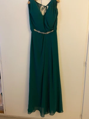 Grön långklänning med pärldetalj - Elegant grön långklänning med v-ringning och pärldetaljer i midjan. Klänningen har omlottliv, knytning i nacken och är tillverkad i ett tunt, flowy material som ger en snygg siluett. Perfekt för bal eller festliga tillfällen.