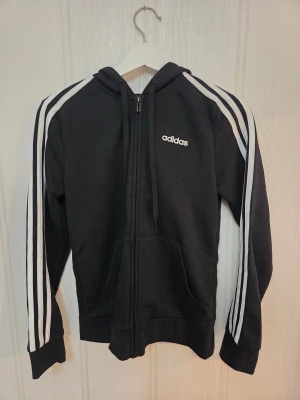 Svart Adidas hoodie med dragkedja - Svart hoodie från Adidas med klassiska vita ränder längs ärmarna och Adidas-logga på bröstet. Tröjan har huva med snörning, dragkedja framtill och en stor ficka på magen. Knappt använd, stl S.