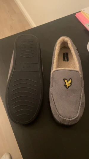 Grå loafers Lyle & Scott - Mysiga grå loafers från Lyle & Scott med mjukt, beige foder och klassisk gul fågel-logga på ovansidan. Skorna har platt sula och rund tå, perfekta för chill hemma. Utsidan är i mocka-look och insidan är fluffig för extra komfort. Använda 1-2 gånger fick dom i present men dom var för små för mig!