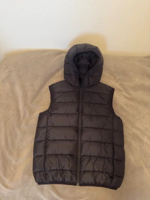 Svart dunväst  H&M - Svart dunväst från H&M med quiltad design och praktisk huva. Västen har dragkedja framtill och är tillverkad i syntetmaterial. Perfekt för lager-på-lager och enkel att matcha med olika outfits.