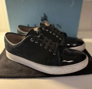 Svarta sneakers från Lanvin med lackad tå - Lanvin sneakers i svart mocka med vit platt sula och snörning. Tåpartiet är i blankt lackskinn som ger en snygg kontrast mot resten av skon. Klassisk rund tå och låg modell, perfekt för dig som gillar stilrena och exklusiva sneakers. Dom har definitivt defekter, där av priset. 