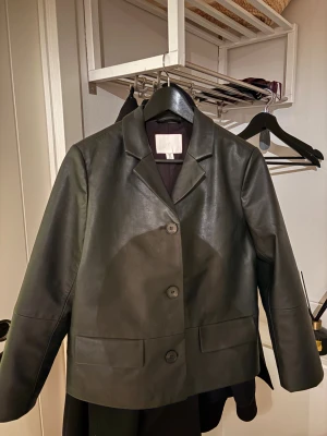 Svart skinnkavaj från H&M - Säljer en svart skinnkavaj från H&M med klassisk krage och tre knappar framtill. Jackan har raka, långa ärmar och två fickor framtill. Perfekt för dig som gillar stilrena och minimalistiska plagg med edge.