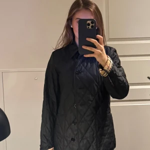 Svart quiltad Burberry jacka - Quiltad svart jacka från Burberry med klassiskt rutigt foder i beige, rött och svart. Jackan har krage, stora knappar framtill och långa ärmar med synlig Burberry-rutig kant. Snygg, tidlös och perfekt för höst och vår. Sparsamt använd, köptes för ca 11 000kr på NK förra året!