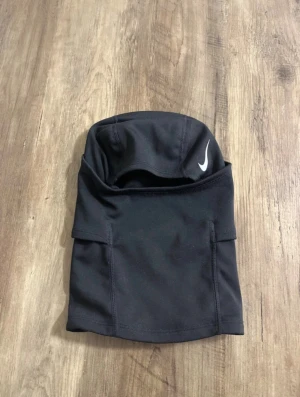 Nike Pro svart ski mask  - Svart Nike Pro mössa/huva i sportig design med vit Nike-logga framtill och etikett bak. Tillverkad i stretchigt syntetmaterial som andas, perfekt för träning utomhus under kyliga dagar. Sitter tight runt huvudet och halsen för extra värme och skydd.
