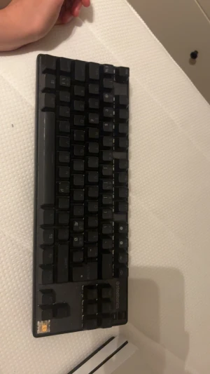 SteelSeries mekaniskt tangentbord - Kompakt SteelSeries mekaniskt tangentbord i svart. Tangenterna ser ut att vara i gott skick, men en tangent saknas i nedre vänstra hörnet. Perfekt för gaming eller arbete, med responsiva mekaniska switchar och robust design.