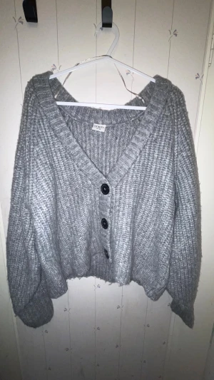 Grå stickad kofta från Stories S - Mysig grå stickad kofta från Stories i storlek S. Koftan har grov ribbad struktur, v-ringad hals och tre svarta knappar framtill. Modellen är lite croppad med långa, breda ärmar och känns perfekt för kyliga dagar.