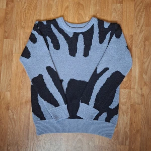 Grå och svart stickad tröja M - Säljer en grå stickad tröja med svart zebramönster i storlek M. Tröjan har rund hals, ribbade muddar. Perfekt för dig som gillar djurmönster och vill ha en unik look.