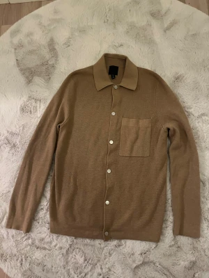 Beige stickad kofta från H&M - Snygg beige stickad kofta från H&M med klassisk krage och silverfärgade knappar framtill. Koftan har en stor bröstficka och långärmade ärmar. Perfekt för lager-på-lager och chill stil. Materialet är mjukt och skönt mot huden.