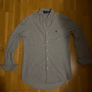 Grå Skjorta Ralph Lauren - Säljer en stilren grå långärmad skjorta från Ralph Lauren i mjuk bomull. Skjortan har klassisk button-down krage, vita knappar och den ikoniska mörkblå broderade loggan på bröstet. 