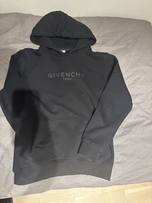 Svart Givenchy hoodie med huva - Svart hoodie från Givenchy med stor logga 'GIVENCHY PARIS' på bröstet. Tröjan har huva, ribbade muddar och en klassisk känguruficka framtill. Materialet är mjukt och känns lyxigt, perfekt för dig som gillar streetwear med designer-vibe.