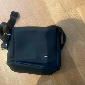 Svart axelväska från Lacoste - Snygg svart axelväska från Lacoste i stilren design med justerbart axelband och diskret krokodillogga i metall på framsidan. Väskan är tillverkad i slätt skinnliknande material och har ett praktiskt lock framtill.