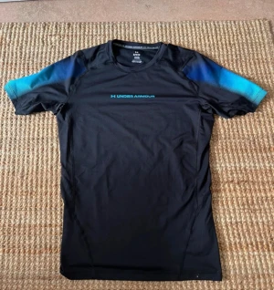 Svart Under Armour tränings-tshirt - Säljer en svart Under Armour HeatGear t-shirt i storlek L. T-shirten har korta ärmar med blå detaljer på axlarna och är tillverkad i ett syntetmaterial som andas. Perfekt för dig som gillar att träna och vill ha en sportig look. Har använts exakt 3 gånger 