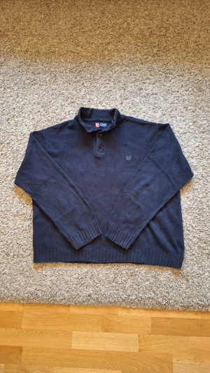 Marinblå stickad tröja från CHAPS med halvpolokrage och knappar - Snygga varm marinblå stickad tröja med halvpolokrage och knappar från CHAPS i storlek XXL, men skulle nog säga den passar mer som XL/L. 100% bomull. Använd fåtal gånger och skicket är riktigt bra, inga defekter eller slitningar finns. Den har en halvpolokrage med knappar. Passar suveränt för dig som gillar stilrena outfits. Skick: 8/10