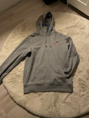 Grå hoodie från GANT med broderi - Snygg grå hoodie från GANT i bomull med broderad logga och emblem på bröstet. Tröjan har huva med snörning, magficka och ribbade muddar. Perfekt för chill dagar och har en klassisk, clean look.