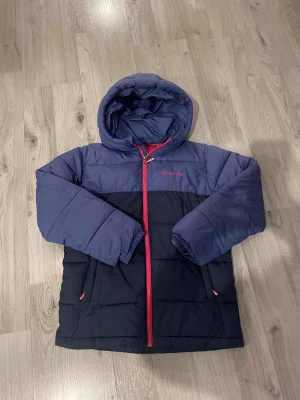 Blå pufferjacka från Columbia - Blå och mörkblå pufferjacka från Columbia med huva och röda dragkedjedetaljer. Jackan har quiltad design, två fickor med dragkedja och logga på bröstet. Perfekt för kalla dagar och har en riktigt skön passform.