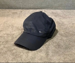 Svart Nike keps Heritage86 - Svart keps från Nike, modell Heritage86, med klassisk böjd skärm och broderad Swoosh-logga framtill. Kepsen är tillverkad i bomull och har justerbar passform baktill. Perfekt för en sportig och avslappnad stil.