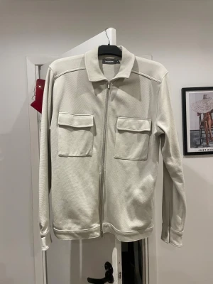 Beige overshirt med dragkedja - Stilren beige overshirt från Dressmann med två stora bröstfickor och dragkedja framtill. Jackan har klassisk krage och långa ärmar med knapp vid ärmslut. Perfekt för lager-på-lager och enkel att matcha till olika outfits.