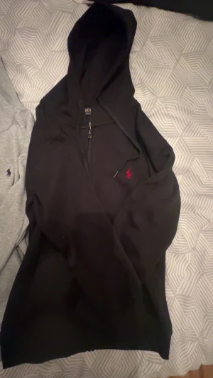 Svart hoodie från Polo Ralph Lauren - Svart hoodie från Polo Ralph Lauren med röd broderad logga på bröstet. Tröjan har huva med snörning och dragkedja framtill. Mjuk bomullskvalitet och långärmad modell som är perfekt för chill dagar.vi kan diskutera om priset.