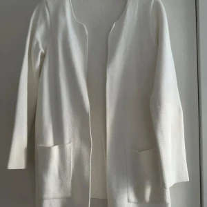 Vit stickad kofta med fickor från Gina Tricot - Säljer en stilren vit kofta med lång ärm och två praktiska fickor framtill. Koftan har en öppen design utan knappar eller dragkedja. Perfekt att slänga över en t-shirt eller topp
