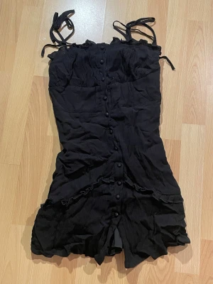 Svart kort klänning från SHEIN - Svart kort klänning från SHEIN med knytbara axelband, volangdetaljer och knappar längs hela framsidan. Klänningen sitter figurnära.