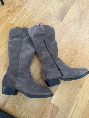 Grå mockastövlar med låg klack - Snygga grå stövlar i mocka med låg klack och dragkedja på insidan. De har en klassisk form med rund tå och diskreta sömdetaljer. Perfekta för dig som vill ha en stilren look med lite western-vibe.