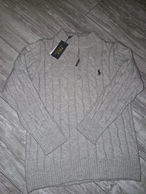 Grå stickad tröja Polo Ralph Lauren Halv zip - Snygg grå kabelstickad tröja från Polo Ralph Lauren med half zip och ribbade muddar. Tröjan har lång ärm och klassisk broderad logga på bröstet. Perfekt för dig som gillar stilren och tidlös design med lite extra edge. Köpt 1300kr.