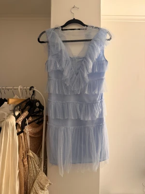 Ljusblå volangklänning i tyll - Supersöt ljusblå kortklänning i tyll med flera lager volanger och v-ringning. Klänningen är ärmlös och har ett diskret prickigt mönster i tyllen. 