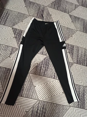 Svarta Adidas träningsbyxor med vita ränder - Säljer ett par svarta Adidas träningsbyxor med klassiska vita ränder längs sidorna och Adidas-logga på benet. Byxorna har elastisk midja med snörning och är gjorda i lätt polyester. Perfekta för sport eller chill. Primegreen-material.