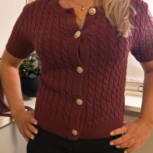 Vinröd kabelstickad kofta med korta ärmar - Säljer en vinröd kabelstickad kofta med korta ärmar och dekorativa guldknappar. Perfekt för en stilren look. Koftan har en klassisk design med ribbade kanter.