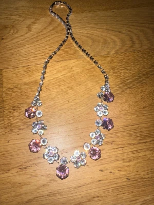 Glittrigt halsband och örhängen med stenar - Säljer ett set med halsband och matchande örhängen i silverfärg med stora ljusrosa och klara stenar. Både halsbandet och örhängena har blommiga former och gnistrar rejält. Perfekt för dig som vill sticka ut med extra bling och färg.
