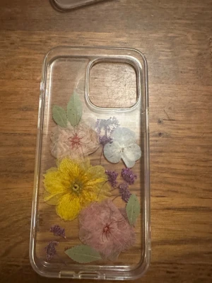Ideal of Sweden Blommigt Skal iPhone - Snyggt genomskinligt mobilskal med pressade blommor från Ideal of Sweden. Passar iPhone-modeller med stor kameramodul. Skalet är i fint skick utan synliga repor eller skador. Perfekt för dig som vill ge din mobil en unik och färgglad look. Till 14 PRO Max 