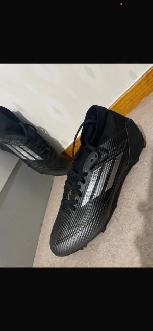 Svarta adidas fotbollsskor med dobbar - Snygga svarta adidas fotbollsskor med vita ränder och gul logga på hälen. Skorna har snörning, platt sula med fasta dobbar och en sportig, strömlinjeformad design. Ovandelen är i syntetmaterial med diskreta linjer för extra stilpoäng. Perfekta för dig som vill sticka ut på planen.