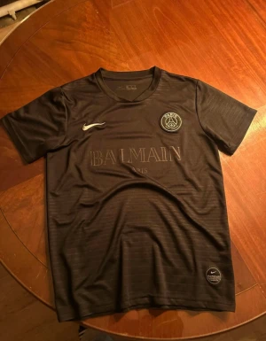 Svart PSG x Balmain fotbollströja Nike - Säljer en svart Paris Saint-Germain x Balmain fotbollströja från Nike. Materialet är lätt och andas, perfekt för träning eller match. PSG-logga och Balmain Paris tryck på bröstet.