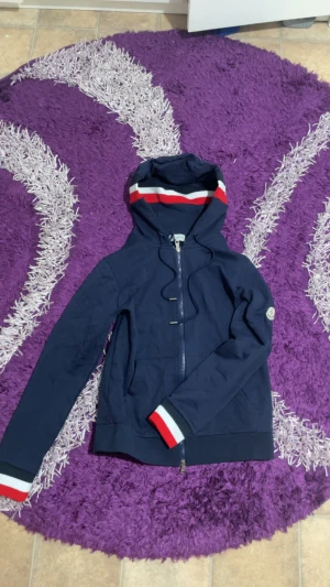 Marinblå vindjacka från Moncler - Snygg marinblå vindjacka från Moncler med huva och dragkedja. Jackan har vita och röda ränder på huva och muddar, samt Moncler-logga på ärmen. Tillverkad i bomull och polyester, perfekt för dig som vill ha en stilren och sportig look.