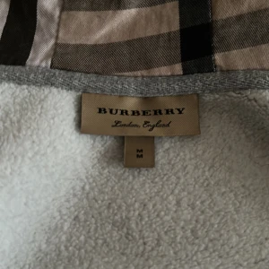 Byte elle köp - Säljer en grå hoodiejacka från Burberry i bomull med klassiskt rutigt foder i beige, svart och rött. Jackan har dragkedja, snörning i huvan och ribbade muddar. Perfekt för dig som vill ha en stilren och lyxig streetwear-look.