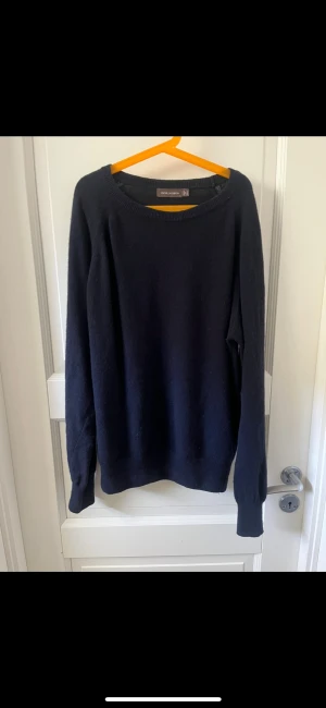 Mörkblå stickad tröja Oscar Jacobson - Mörkblå stickad tröja från Oscar Jacobson, tillverkad i Italien. Tröjan har rund halsringning, ribbade muddar och lång ärm. Enkel och stilren design som funkar till många olika looks. Perfekt för dig som gillar klassiska plagg med snygg passform. Tröjan har en liten fläck på armen. Går o tvätta bort.