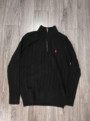 Svart stickad tröja Polo Ralph Lauren - Svart kabelstickad tröja från Polo Ralph Lauren med halv dragkedja och ribbad krage. Klassisk röd logga broderad på bröstet. Tröjan har långärmad passform och är tillverkad i mjuk bomull, perfekt för dig som gillar stilren och tidlös design.