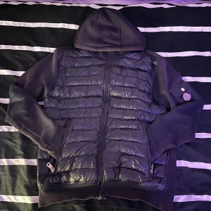 Svart pufferjacka med huva från Moncler - Svart cardigan från Moncler med huva och quiltad framsida. Jackan har släta ärmar, dragkedja framtill och två fickor med dragkedja. Moncler-logga på ärmen och en liten ficka med knapp. Perfekt för kyliga dagar. Den börjar tappa lite färg på luvan och ärmarna men är väldigt fin. Priset kan diskuteras. Samt ärmarna är lite skadade men ska sys ihop. 