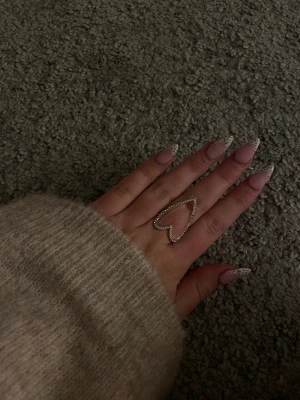 Silverring med hjärtform och stenar - Superfin silverfärgad ring i form av ett öppet hjärta, dekorerad med små glittrande stenar runt hela hjärtat. Perfekt statement-piece för dig som gillar unika smycken och vill sticka ut lite extra.