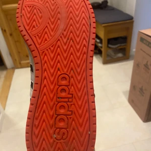 Svarta adidas sneakers med röda detaljer - Snygga svarta adidas sneakers med högt skaft, vita ränder på sidorna och röd insida. Yttersulan är röd med tydligt adidas-tryck. Skorna har snörning och är tillverkade i syntet och textil. Perfekta för dig som gillar sportig stil och vill sticka ut lite extra. När skick måste bara tvättas