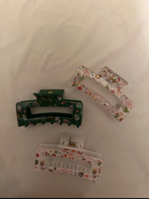 Juliga hårklämmor med motiv - Tre hårklämmor i plast med juliga motiv som tomtar, snögubbar och julgranar. En är grön med mönster, två är vita med färgglada detaljer. Perfekt för att ge din frisyr en festlig touch under vintern och julen.